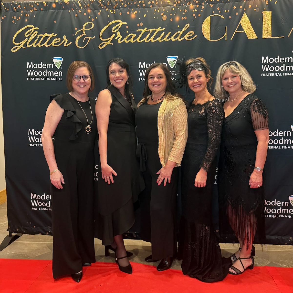 Five women stand together, smiling, on a red carpet in front of a black backdrop at a formal event. They are all dressed in elegant black evening attire, including dresses and jumpsuits. The woman in the center wears a shimmering gold cardigan over her black outfit, and the woman to her right wears a delicate black lace headpiece. The backdrop features the words "Glitter & Gratitude GALA" in large gold script at the top, along with repeated white logos for "Modern Woodmen FRATERNAL FINANCIAL.