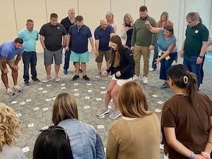 A high-angle shot of a large group of adults participating in a team-building activity or icebreaker game. They are standing in a wide circle around a woman in a black jacket and white shorts who is stepping on small, numbered white cards scattered across the carpeted floor. The participants, dressed in business-casual attire like polos, button-downs, and t-shirts, are leaning forward and pointing with focused, smiling expressions. The room has neutral-toned walls and professional lighting.