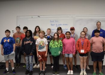 A diverse group of thirteen children and one adult male stand together for a group photo in front of a large whiteboard in what appears to be a classroom or community center. The children, a mix of boys and girls of various ages, are dressed in casual clothing like t-shirts, hoodies, and athletic wear. Some wear hats or lanyards. The whiteboard behind them has "Ground Rules" written at the top in blue marker, followed by a list of expectations. The man on the far right is smiling and wearing a blue polo shirt with a small logo. The room has neutral gray walls and a dark gray carpeted floor