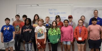 A diverse group of thirteen children and one adult male stand together for a group photo in front of a large whiteboard in what appears to be a classroom or community center. The children, a mix of boys and girls of various ages, are dressed in casual clothing like t-shirts, hoodies, and athletic wear. Some wear hats or lanyards. The whiteboard behind them has "Ground Rules" written at the top in blue marker, followed by a list of expectations. The man on the far right is smiling and wearing a blue polo shirt with a small logo. The room has neutral gray walls and a dark gray carpeted floor