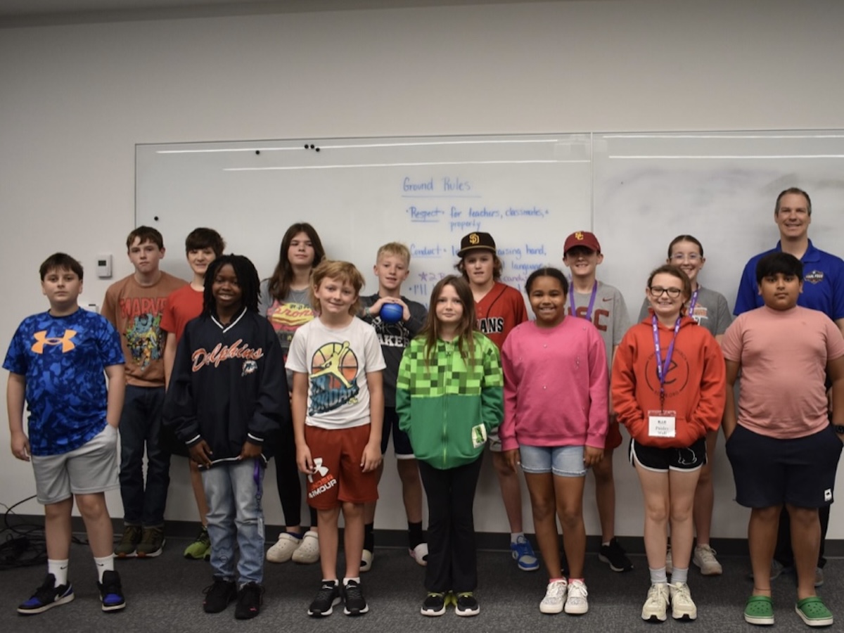A diverse group of thirteen children and one adult male stand together for a group photo in front of a large whiteboard in what appears to be a classroom or community center. The children, a mix of boys and girls of various ages, are dressed in casual clothing like t-shirts, hoodies, and athletic wear. Some wear hats or lanyards. The whiteboard behind them has "Ground Rules" written at the top in blue marker, followed by a list of expectations. The man on the far right is smiling and wearing a blue polo shirt with a small logo. The room has neutral gray walls and a dark gray carpeted floor