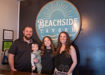 A family of four stands together for a professional photo in front of a prominent circular logo for Beachside Tavern. The family includes a man with a beard, two women, and a toddler. The tavern's teal and gold logo, which features a sun and wave design, is set against a dark blue accent wall. The group is smiling and appears to be in a welcoming, indoor restaurant setting.