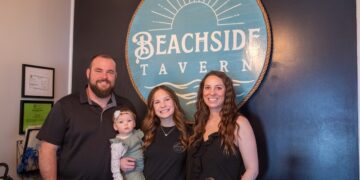 A family of four stands together for a professional photo in front of a prominent circular logo for Beachside Tavern. The family includes a man with a beard, two women, and a toddler. The tavern's teal and gold logo, which features a sun and wave design, is set against a dark blue accent wall. The group is smiling and appears to be in a welcoming, indoor restaurant setting.