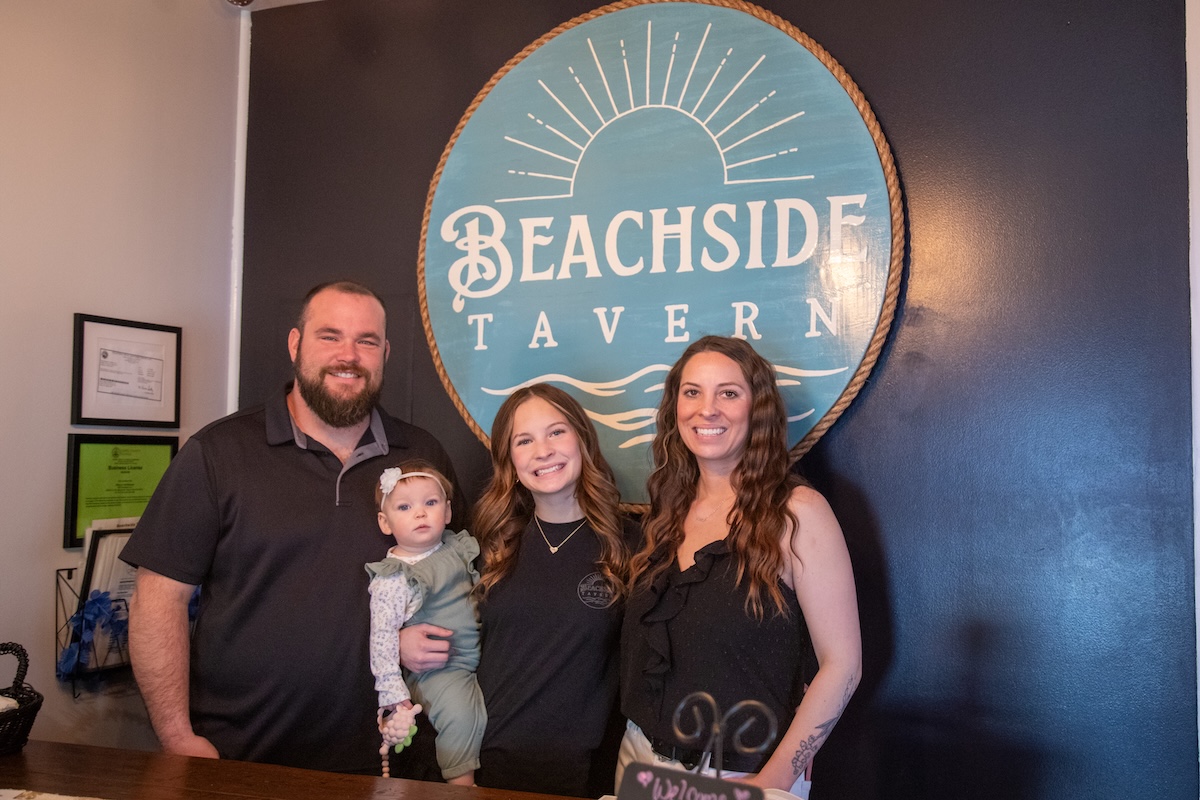 A family of four stands together for a professional photo in front of a prominent circular logo for Beachside Tavern. The family includes a man with a beard, two women, and a toddler. The tavern's teal and gold logo, which features a sun and wave design, is set against a dark blue accent wall. The group is smiling and appears to be in a welcoming, indoor restaurant setting.