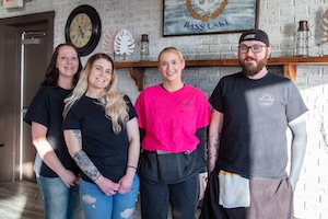 Four staff members of Beachside Tavern stand together and smile for a group photo in front of a rustic grey-brick wall. The three women and one man are dressed in casual professional attire, including black crewneck t-shirts and a vibrant pink scrub-style top. The background features a wooden mantel decorated with a white leaf sculpture and a framed sign that says "Bass Lake," highlighting the restaurant's location.