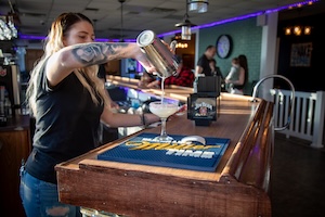 A professional bartender with a floral arm tattoo is seen in action at Beachside Tavern, pouring a freshly shaken cocktail from a stainless steel shaker into a chilled coupe glass. The scene is set at the tavern's polished wood bar, featuring a prominent blue and gold "Miller Time" bar mat. In the blurred background, patrons sit at the bar under modern industrial pendant lights and vibrant purple ambient lighting.