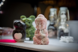 A close-up, slightly blurred shot of several small decorative figurines on a white surface. The central focus is a small, hand-carved pink opal Buddha in a seated meditation pose. Behind it, a green flocked moss bunny and a black heart-shaped figurine are visible. In the background, two glass jars with white stoppers are softly out of focus, all set against a dark backdrop with warm string lights.