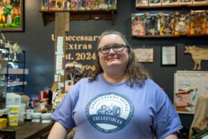 A woman with wavy brown hair and green-rimmed glasses is smiling in front of a dark grey wall in a boutique setting. She is wearing a light purple t-shirt featuring the logo for Unnecessary Extras Collectibles, which includes a classic tractor design. Behind her, shelves are filled with a large collection of boxed Barbie and The Wizard of Oz collector dolls, and a sign on the wall reads "Unnecessary Extras Est. 2024."