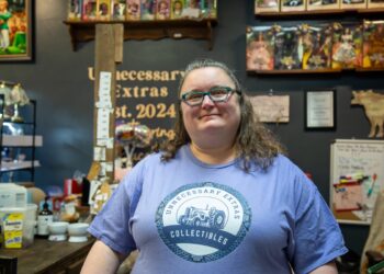 A woman with wavy brown hair and green-rimmed glasses is smiling in front of a dark grey wall in a boutique setting. She is wearing a light purple t-shirt featuring the logo for Unnecessary Extras Collectibles, which includes a classic tractor design. Behind her, shelves are filled with a large collection of boxed Barbie and The Wizard of Oz collector dolls, and a sign on the wall reads "Unnecessary Extras Est. 2024."