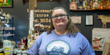 A woman with wavy brown hair and green-rimmed glasses is smiling in front of a dark grey wall in a boutique setting. She is wearing a light purple t-shirt featuring the logo for Unnecessary Extras Collectibles, which includes a classic tractor design. Behind her, shelves are filled with a large collection of boxed Barbie and The Wizard of Oz collector dolls, and a sign on the wall reads "Unnecessary Extras Est. 2024."
