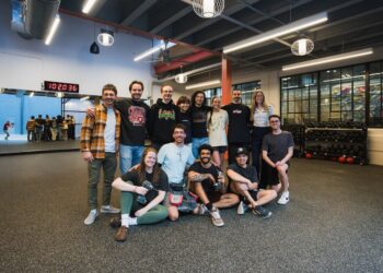 A diverse group of thirteen smiling people pose for a group photo inside a modern, industrial-style climbing gym or fitness center. The group is arranged in two rows, with nine people standing and four people sitting or kneeling on the dark gray textured floor. They are dressed in casual athletic wear, including t-shirts, hoodies, and flannels. In the background, a large mirror reflects more gym-goers, and a digital clock displays "10:20:36." To the right, racks of kettlebells and a climbing wall are visible behind a glass partition. The space is brightly lit by large rectangular overhead lights and spherical pendant lamps.