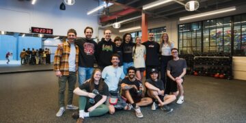 A diverse group of thirteen smiling people pose for a group photo inside a modern, industrial-style climbing gym or fitness center. The group is arranged in two rows, with nine people standing and four people sitting or kneeling on the dark gray textured floor. They are dressed in casual athletic wear, including t-shirts, hoodies, and flannels. In the background, a large mirror reflects more gym-goers, and a digital clock displays "10:20:36." To the right, racks of kettlebells and a climbing wall are visible behind a glass partition. The space is brightly lit by large rectangular overhead lights and spherical pendant lamps.