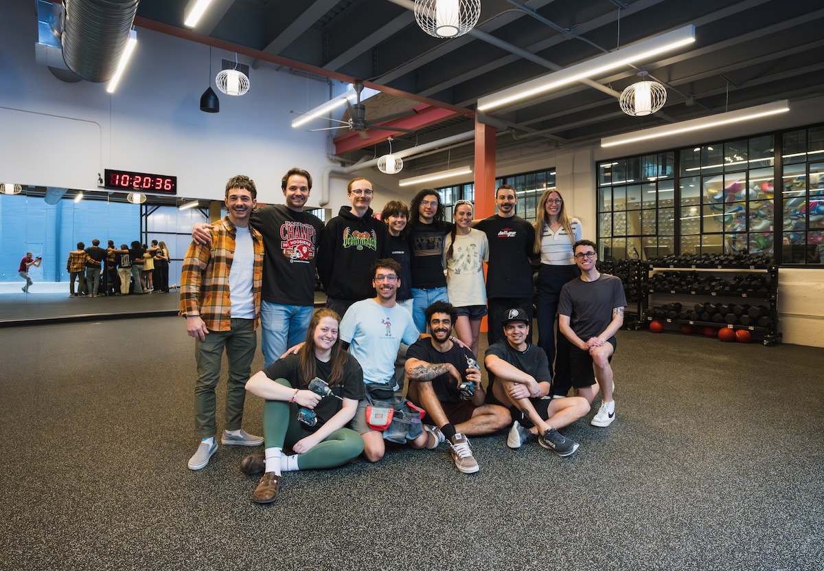 A diverse group of thirteen smiling people pose for a group photo inside a modern, industrial-style climbing gym or fitness center. The group is arranged in two rows, with nine people standing and four people sitting or kneeling on the dark gray textured floor. They are dressed in casual athletic wear, including t-shirts, hoodies, and flannels. In the background, a large mirror reflects more gym-goers, and a digital clock displays "10:20:36." To the right, racks of kettlebells and a climbing wall are visible behind a glass partition. The space is brightly lit by large rectangular overhead lights and spherical pendant lamps.