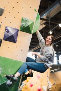 A smiling young woman with dark hair pulled back is indoor bouldering. She is wearing a gray long-sleeved sweatshirt, dark teal leggings, and black climbing shoes. She is positioned on a light-colored wooden climbing wall, hanging from a large, circular green hold with her right hand while her left arm is bent. Her left foot is braced against a small gray hold on a green section of the wall. Several other geometric holds in green, purple, and gray are visible. The background shows the industrial ceiling of the gym with exposed pipes and lights.