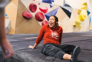 A woman with long dark hair sits on a thick, dark gray crash pad in a brightly lit indoor bouldering gym. She is laughing joyfully with her mouth open, looking toward someone off-camera to her left. She is wearing a long-sleeved red shirt and dark cargo pants. In the soft-focus background, a wooden climbing wall is covered in various bouldering holds, including large red spheres and angular yellow shapes.