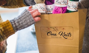A close-up shot focuses on a brown paper shopping bag from "Oma Knits Textile Co." held by a person wearing a yellow and brown patterned sleeve and a gray ribbed fingerless glove. Inside the bag, several skeins of vibrant yarn in shades of cream, purple, bright pink, and forest green are visible, each with a white "Oma Knits" label. The bag is placed on a light-colored counter in a brightly lit shop with a soft-focus background.