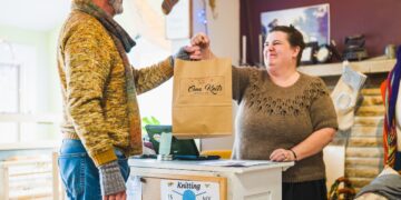 A medium shot in a brightly lit, cozy yarn shop shows a woman with dark hair pulled back, wearing a brown textured knit sweater, standing behind a white wooden counter and smiling warmly as she hands a brown paper shopping bag to a man. The man, who has a gray beard and glasses, is dressed in a yellow and orange knit sweater and gray fingerless gloves. The shopping bag features the logo "Oma Knits Textile Co." and the counter has a small corkboard with a sign that reads "Knitting is my superpower" with a blue ball of yarn illustration. The background is filled with various knitting-related items, including stockings hanging on the wall and colorful yarn.