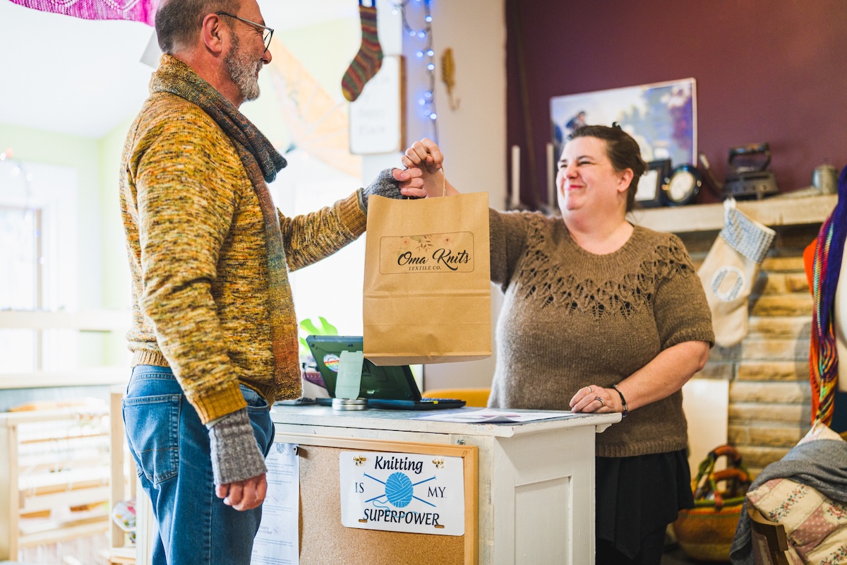 A medium shot in a brightly lit, cozy yarn shop shows a woman with dark hair pulled back, wearing a brown textured knit sweater, standing behind a white wooden counter and smiling warmly as she hands a brown paper shopping bag to a man. The man, who has a gray beard and glasses, is dressed in a yellow and orange knit sweater and gray fingerless gloves. The shopping bag features the logo "Oma Knits Textile Co." and the counter has a small corkboard with a sign that reads "Knitting is my superpower" with a blue ball of yarn illustration. The background is filled with various knitting-related items, including stockings hanging on the wall and colorful yarn.