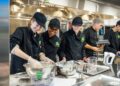 Three smiling young women and two young men work together in a professional commercial kitchen at Westfield High School. The students are dressed in black chef's coats with a green shamrock logo and black baseball caps or hairnets. In the foreground, the women are mixing ingredients in large stainless steel bowls on a metal counter scattered with flour. Various kitchen tools, including measuring cups, a whisk, and a hand mixer, are spread across the workspace. The background features industrial stainless steel ovens and shelving under bright fluorescent lights.
