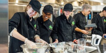 Three smiling young women and two young men work together in a professional commercial kitchen at Westfield High School. The students are dressed in black chef's coats with a green shamrock logo and black baseball caps or hairnets. In the foreground, the women are mixing ingredients in large stainless steel bowls on a metal counter scattered with flour. Various kitchen tools, including measuring cups, a whisk, and a hand mixer, are spread across the workspace. The background features industrial stainless steel ovens and shelving under bright fluorescent lights.