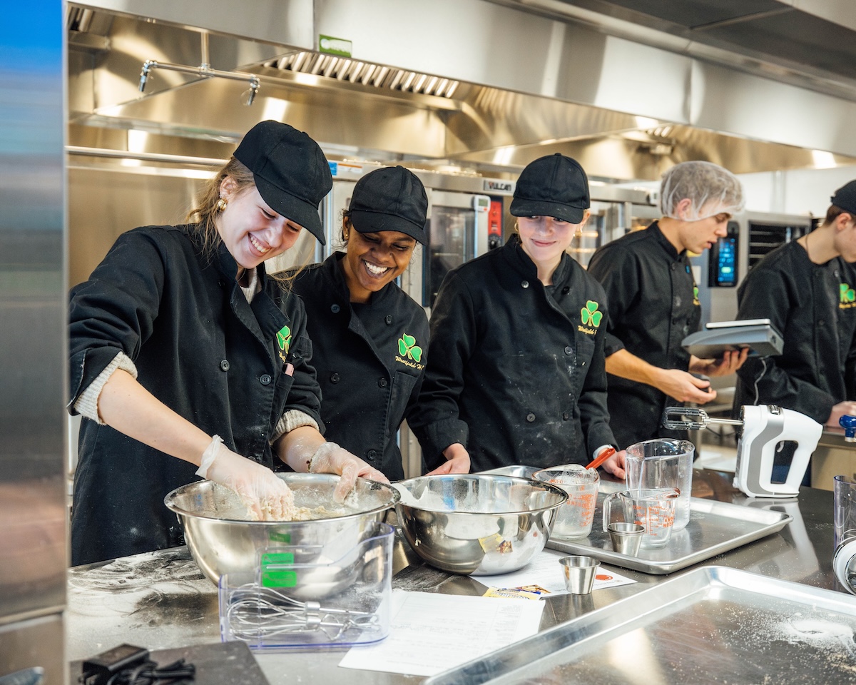 Three smiling young women and two young men work together in a professional commercial kitchen at Westfield High School. The students are dressed in black chef's coats with a green shamrock logo and black baseball caps or hairnets. In the foreground, the women are mixing ingredients in large stainless steel bowls on a metal counter scattered with flour. Various kitchen tools, including measuring cups, a whisk, and a hand mixer, are spread across the workspace. The background features industrial stainless steel ovens and shelving under bright fluorescent lights.