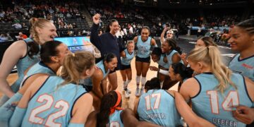A wide-angle, low-perspective shot captures a joyful huddle of the Indy Ignite professional women's volleyball team on the court at the Fishers Event Center. The players, wearing light blue and dark blue "Ignite" jerseys, are gathered in a tight circle, laughing and celebrating with their arms around each other. Visible jersey numbers include 22 (Lydia Martyn), 14 (Anna DeBeer), and 17 (Blake Mohler). A coach in a dark navy tracksuit stands in the center of the huddle, smiling and holding up a small object. The background shows a large, blurred crowd in the arena stands under bright stadium lights.
