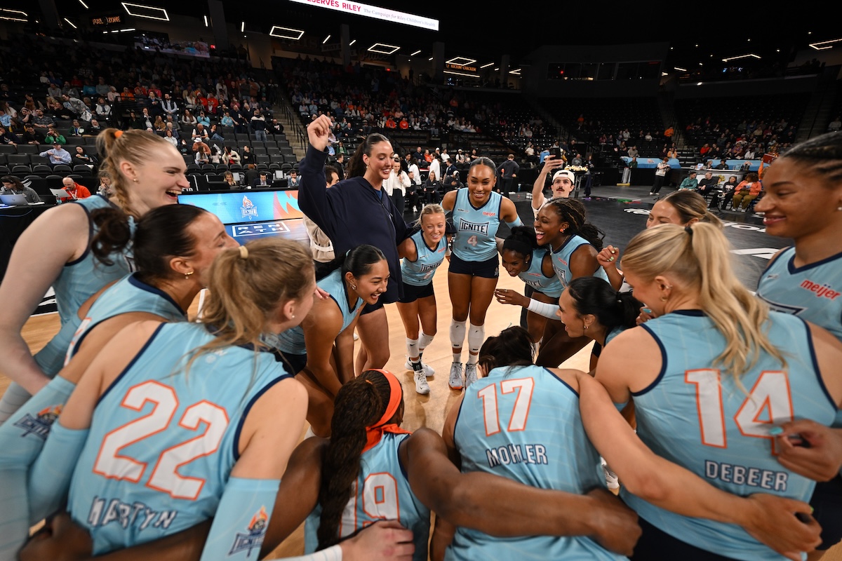 A wide-angle, low-perspective shot captures a joyful huddle of the Indy Ignite professional women's volleyball team on the court at the Fishers Event Center. The players, wearing light blue and dark blue "Ignite" jerseys, are gathered in a tight circle, laughing and celebrating with their arms around each other. Visible jersey numbers include 22 (Lydia Martyn), 14 (Anna DeBeer), and 17 (Blake Mohler). A coach in a dark navy tracksuit stands in the center of the huddle, smiling and holding up a small object. The background shows a large, blurred crowd in the arena stands under bright stadium lights.