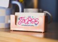 A close-up, slightly angled shot features a small stack of business cards in a glossy, peach-colored plastic holder resting on a light wood surface. The top card features the logo for "THE SOURCE" in a playful, vibrant pink cursive font, accented with small blue and pink starbursts. Beneath the name, the words "curated artistry" are written in a delicate, dark blue script. In the blurred background, a blue and white checkered mug and other desktop items are visible.