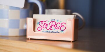A close-up, slightly angled shot features a small stack of business cards in a glossy, peach-colored plastic holder resting on a light wood surface. The top card features the logo for "THE SOURCE" in a playful, vibrant pink cursive font, accented with small blue and pink starbursts. Beneath the name, the words "curated artistry" are written in a delicate, dark blue script. In the blurred background, a blue and white checkered mug and other desktop items are visible.