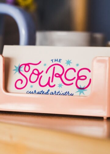 A close-up, slightly angled shot features a small stack of business cards in a glossy, peach-colored plastic holder resting on a light wood surface. The top card features the logo for "THE SOURCE" in a playful, vibrant pink cursive font, accented with small blue and pink starbursts. Beneath the name, the words "curated artistry" are written in a delicate, dark blue script. In the blurred background, a blue and white checkered mug and other desktop items are visible.