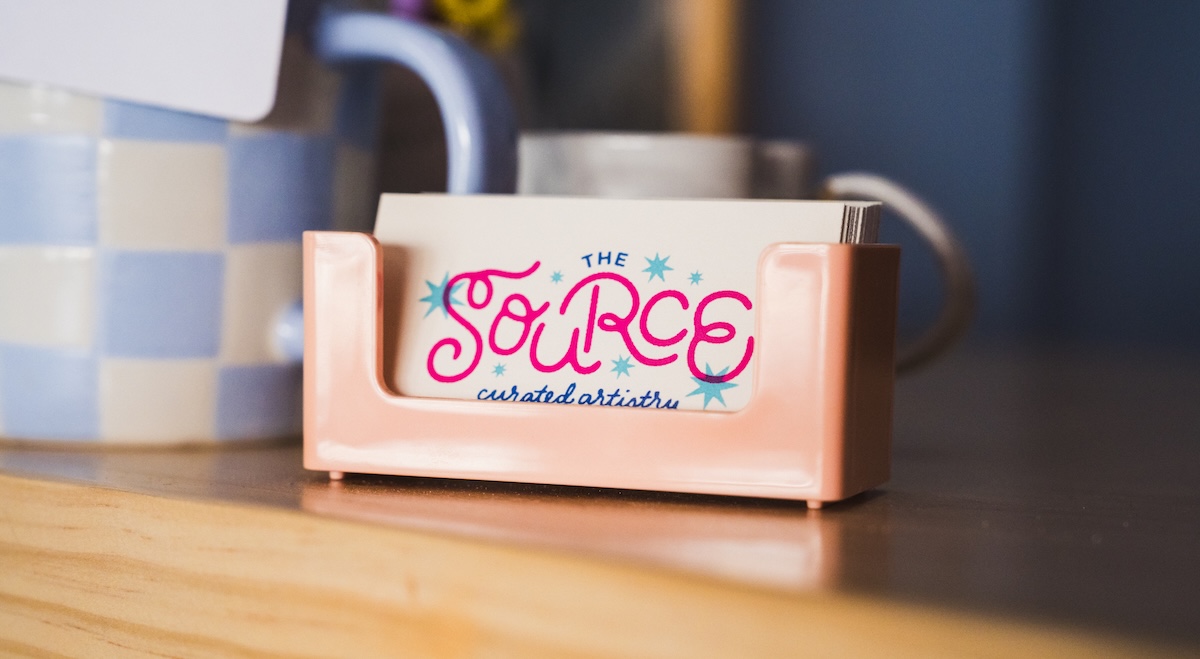 A close-up, slightly angled shot features a small stack of business cards in a glossy, peach-colored plastic holder resting on a light wood surface. The top card features the logo for "THE SOURCE" in a playful, vibrant pink cursive font, accented with small blue and pink starbursts. Beneath the name, the words "curated artistry" are written in a delicate, dark blue script. In the blurred background, a blue and white checkered mug and other desktop items are visible.