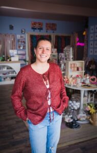 A smiling young woman with her hair pulled back stands in the center of a brightly decorated boutique. She is wearing a maroon, tie-front cardigan over a white top and light-wash ripped jeans. The background is a cozy shop called The Source, featuring blue walls, wooden floors, and various shelves and tables filled with colorful curated gifts, jewelry, and local artwork. A neon pink sign glows faintly in the blurred background.
