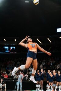 A dynamic, low-angle action shot of Indy Ignite setter Mia Tuaniga (#91) mid-air during a volleyball match. She is jumping high, her body arched as she prepares to set or "dump" the volleyball, which is visible at the top of the frame. She wears the team's orange and blue jersey, dark blue shorts, white knee pads, and white athletic shoes. In the background, teammates in navy tracksuits watch from the sideline, and a large, blurred crowd fills the arena seats. Overhead, linear stadium lights illuminate the court.