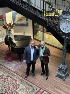 An elevated, high-angle shot looks down into the elegant, light-filled lobby of the Bellasera Resort in Naples, Florida. Two men stand together on the polished wood floor at the base of a grand, dark-wood staircase with a decorative patterned carpet runner. One man wears a navy blue blazer, and the other wears a brown jacket with a light green scarf.The lobby is beautifully appointed with a black grand piano, a large traditional area rug, and an ornate, vintage-style post clock. In the background, a glass-walled gallery reveals a classic red car on display.