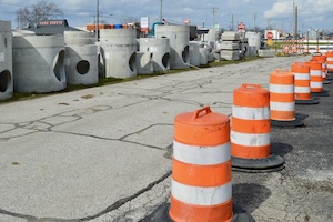A row of bright orange and white construction barrels lines a cracked asphalt area, guiding traffic away from a storage site. Behind the barrels, dozens of large, grey concrete storm drain segments and manhole rings are staged for an upcoming project. The background shows a commercial area under a bright, partly cloudy sky.