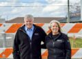 A smiling man and woman stand together outdoors behind a bright orange and white striped construction barricade. Both are wearing matching black zip-up fleece jackets with a small white logo on the left chest that reads "GENUINE FRANKLIN." The man on the left has grey hair and a mustache, wearing a blue checkered button-down shirt under his fleece. The woman on the right has blonde hair and is wearing a black top. The background shows a construction site with concrete structures, power lines, and the "PANA DONUTS" sign in the distance under a cloudy sky.