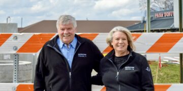 A smiling man and woman stand together outdoors behind a bright orange and white striped construction barricade. Both are wearing matching black zip-up fleece jackets with a small white logo on the left chest that reads "GENUINE FRANKLIN." The man on the left has grey hair and a mustache, wearing a blue checkered button-down shirt under his fleece. The woman on the right has blonde hair and is wearing a black top. The background shows a construction site with concrete structures, power lines, and the "PANA DONUTS" sign in the distance under a cloudy sky.