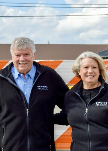 A smiling man and woman stand together outdoors behind a bright orange and white striped construction barricade. Both are wearing matching black zip-up fleece jackets with a small white logo on the left chest that reads "GENUINE FRANKLIN." The man on the left has grey hair and a mustache, wearing a blue checkered button-down shirt under his fleece. The woman on the right has blonde hair and is wearing a black top. The background shows a construction site with concrete structures, power lines, and the "PANA DONUTS" sign in the distance under a cloudy sky.
