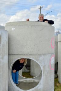 A playful photo features a man and a woman posing with large, precast concrete manhole structures in an outdoor storage yard. A smiling woman in a dark blue sweater and bright blue pants peeks through a large, circular opening at the base of one of the grey concrete cylinders. Above her, a man in a black jacket leans over the top rim of the same structure, smiling warmly at the camera. The background is filled with several more of these heavy industrial concrete forms under a bright, cloud-filled sky. 