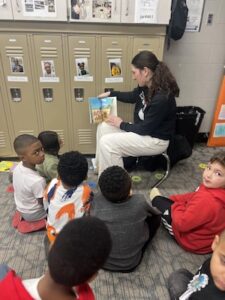 A young woman wearing a long-sleeved navy Lawrence North athletic shirt and white pants sits on a small chair in a classroom, reading a picture book to a group of young children. She is holding the book open, showing an illustration to six children who are sitting on a grey carpeted floor in front of her. In the background, a row of tan school lockers is visible, each featuring a small photo of a student above the vent. The room has a beige wall and various classroom posters.