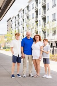A group portrait featuring a man in a blue polo, a woman in a white short-sleeved romper, and two boys in casual summer attire standing on a paved walkway.