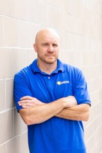 Medium shot of a man in a royal blue polo shirt with a small yellow and white logo, standing outdoors against a grey masonry wall with his arms folded.
