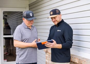 Two men standing outside a house with white siding; one man in a black pullover shows the other, wearing a grey polo and an Indiana University hat, information on a blue tablet in a rugged keyboard case.