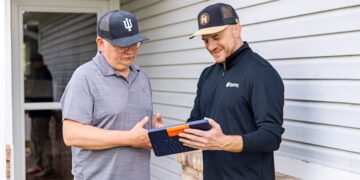Two men standing outside a house with white siding; one man in a black pullover shows the other, wearing a grey polo and an Indiana University hat, information on a blue tablet in a rugged keyboard case.