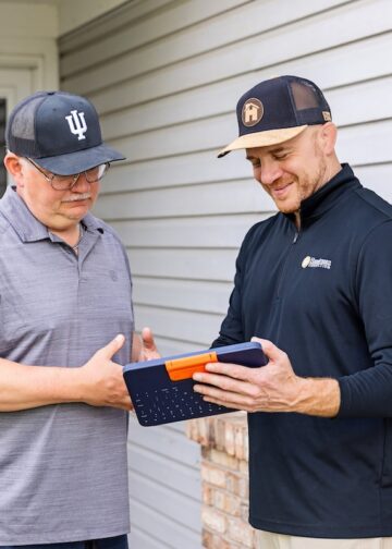 Two men standing outside a house with white siding; one man in a black pullover shows the other, wearing a grey polo and an Indiana University hat, information on a blue tablet in a rugged keyboard case.