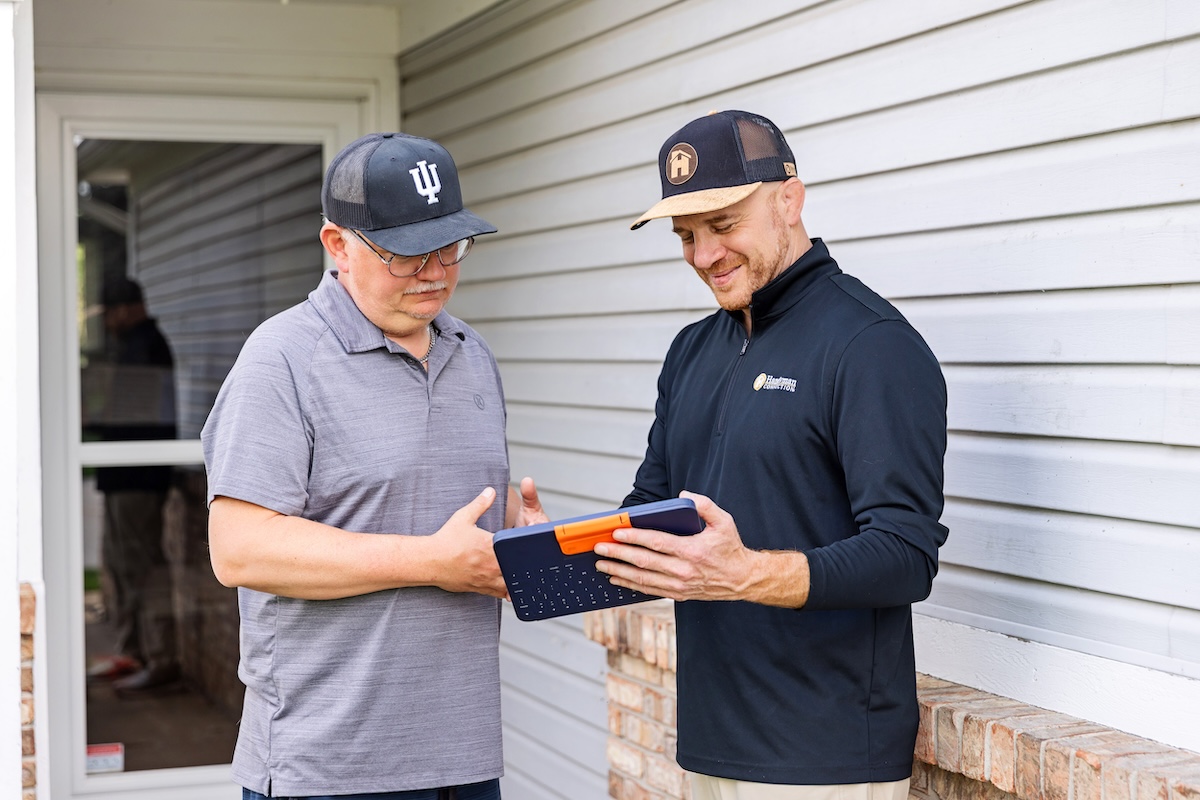 Two men standing outside a house with white siding; one man in a black pullover shows the other, wearing a grey polo and an Indiana University hat, information on a blue tablet in a rugged keyboard case.