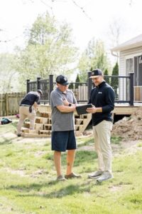 Full-length shot of two men reviewing information on a tablet in a backyard while construction on a new deck and staircase continues behind them.