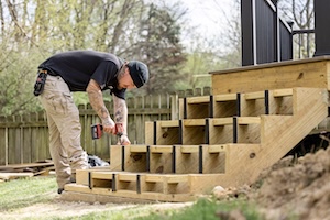 Side view of a man with tattoos on his arms working on a multi-level wooden staircase frame in a backyard, with a wooden fence and trees in the background.