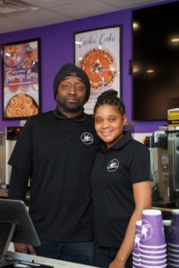 A man and a woman stand close together behind a bakery service counter, both smiling at the camera. They are wearing matching black polo shirts with the white circular Cinnaholic logo. The man is also wearing a black beanie. The background features bright purple walls decorated with framed posters for a "Toppings Bar" and a "Cookie Cake." A stack of purple branded cups sits on the counter in the foreground.