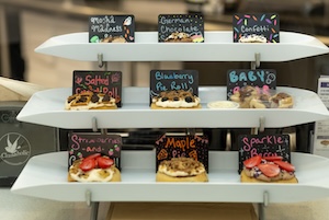 A three-tiered white serving stand displays nine gourmet cinnamon rolls, each with its own decorative chalkboard label. The flavors shown include "Mocha Madness," "German Chocolate," "Confetti," "Salted Caramel," "Blueberry Pie," "Baby," "Strawberry," "Maple Pie," and "Sparkle." The rolls are intricately topped with various frostings, fruits, and crumbles, arranged in a professional bakery setting.
